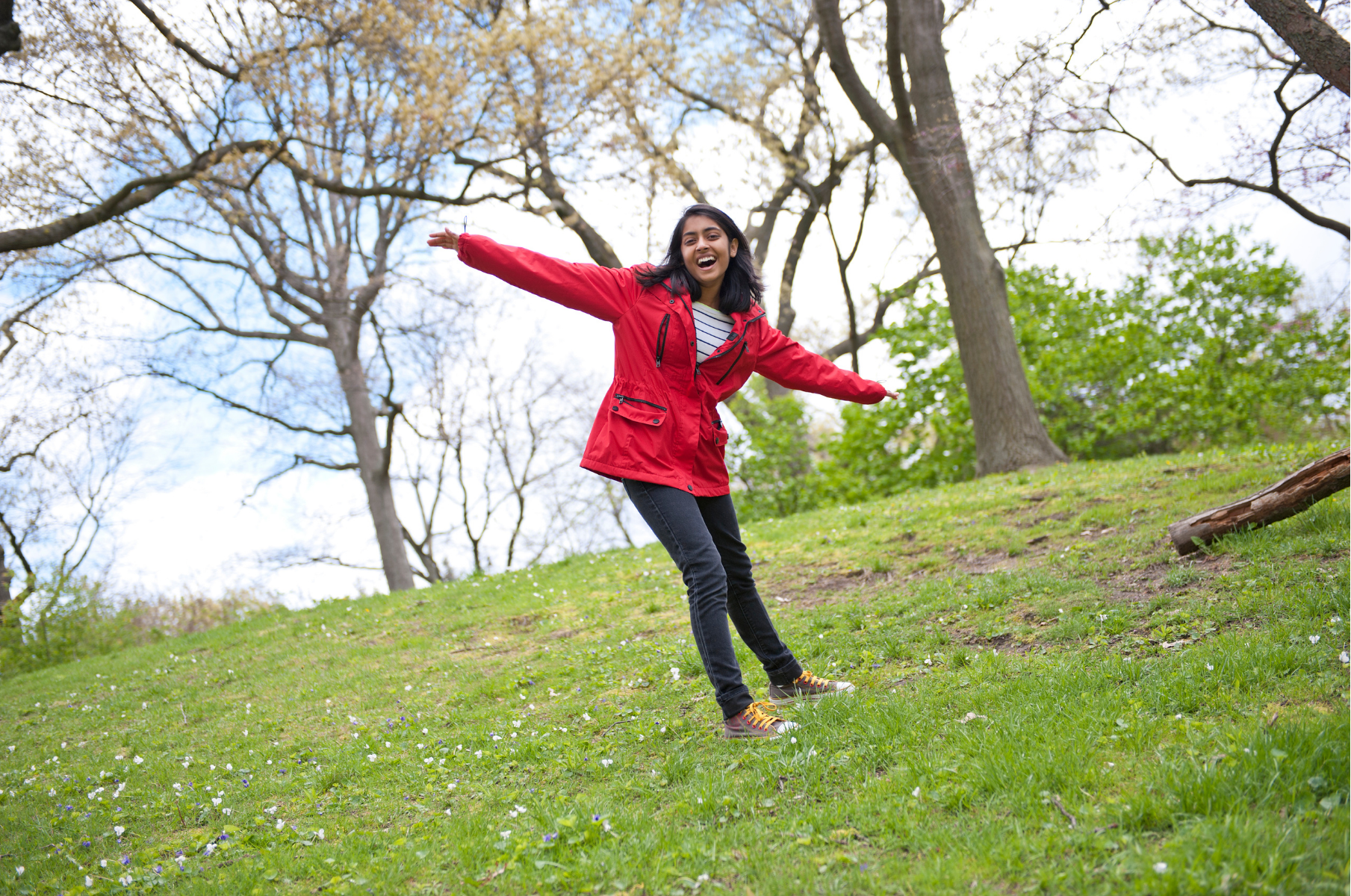 A tween or teen girl with dark hair and skin stands on a grassy hill wearing a red coat and jeans and extending her arms wide in gratitude.