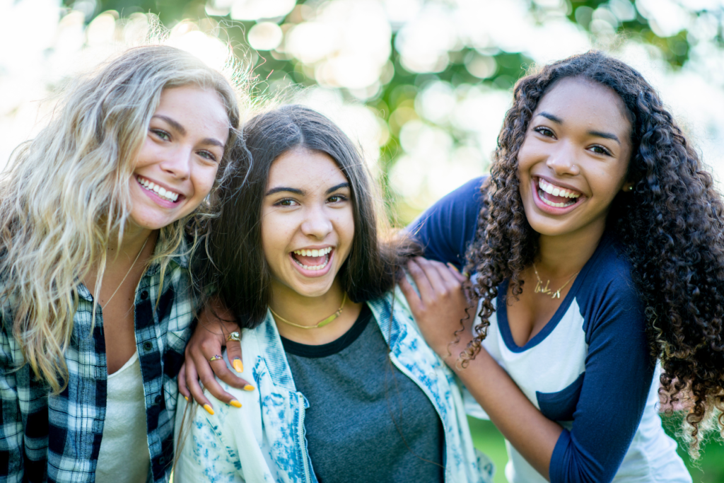 Three tween or teen girls in casual cool-toned clothes laugh together with blurred trees in the background.