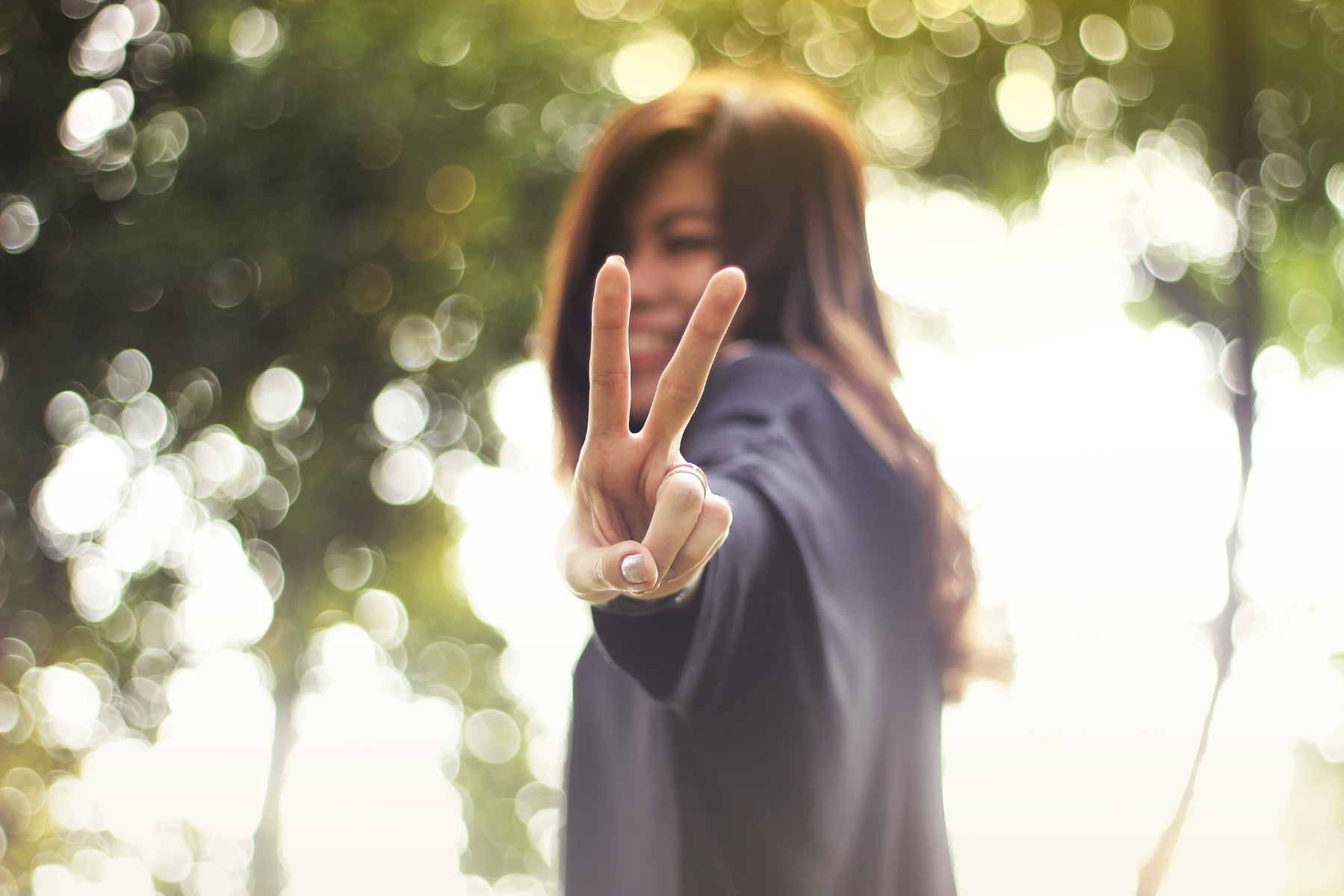 A tween or teen girl with brown hair and skin wearing a gray shirt stands outside near trees holding up a peace sign to the camera.