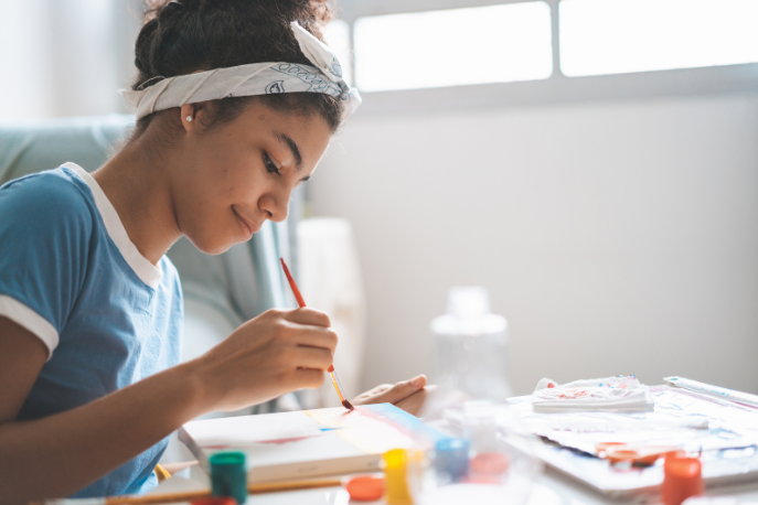 A tween or teen girl with tan skin and brown hair tied up in a bun sits at a table and paints a canvas.