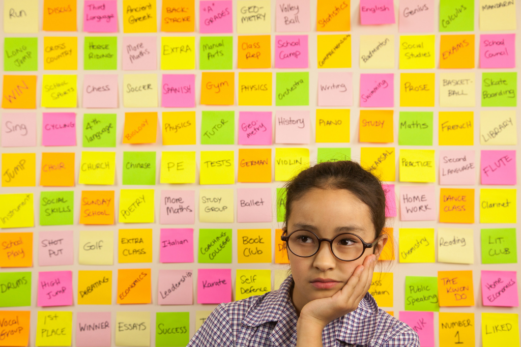 Teen or Tween girl wearing glasses holds her head in her hand overwhelmed by the wall covered in sticky notes behind her that list all of the things to do.