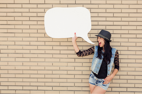 Teen or tween girl with long brown hair wearing a gray hat, black shirt, jean vest and shorts stands next to a tan brick wall holding a blank speech bubble poster