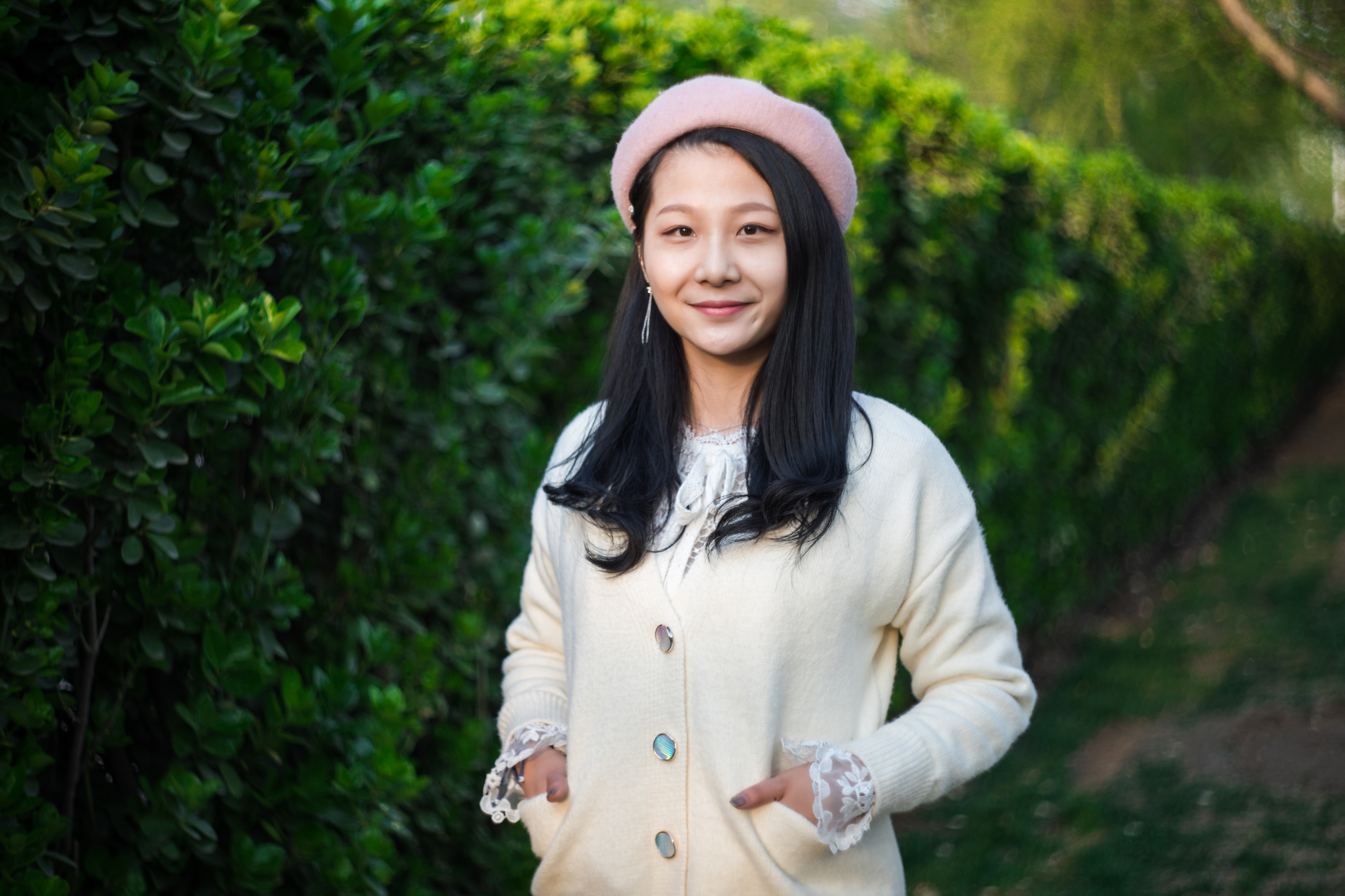 A tween or teen girl with dark hair and tan skin stands in front of tall green bushes wearing a pink beret and cream sweater and smiles practicing affirmations.