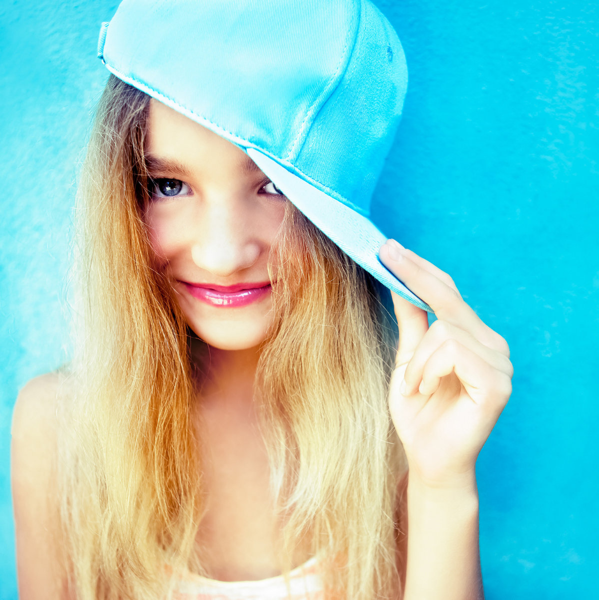 A tween or teen girl with light skin and hair wearing a light blue hat stands in front of a light blue wall and smiles knowing she is unique.