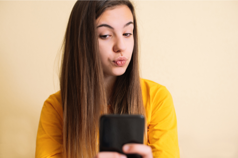 A tween or teen girl with brown hair and a yellow shirt poses with a kissy face to take a selfie with a filter.