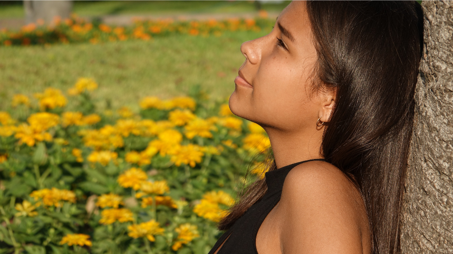 A tween or teen girl with dark hair and tan skin wearing a black tank top rests against a tree while sitting in a field of yellow flowers.