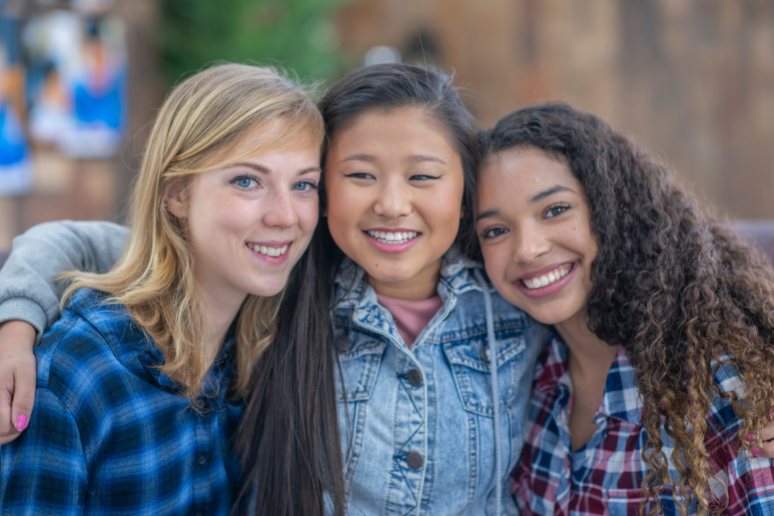 Three tween or Teen girls hug together for a group picture, one has fair skin and blonde hair, and two have tan skin and brown hair.