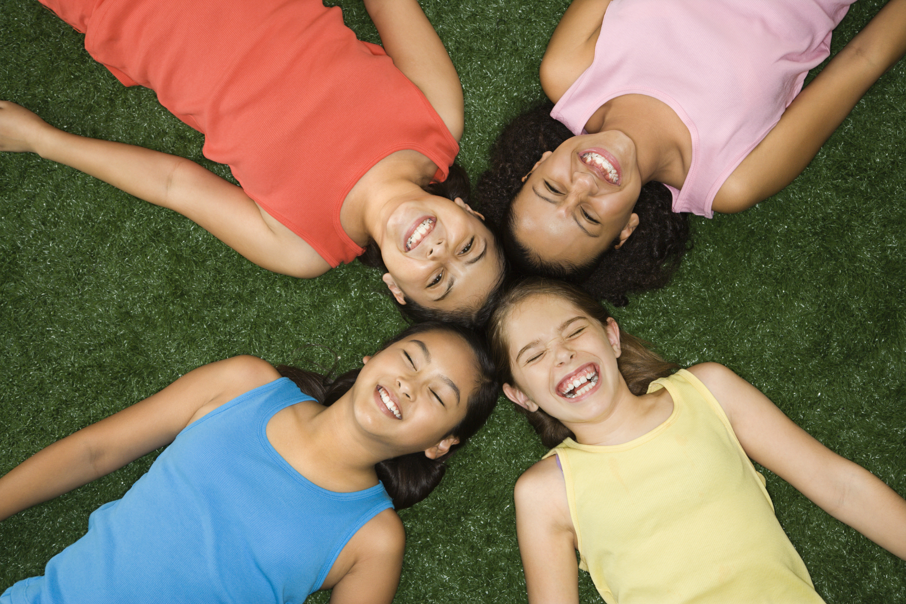 Four tween or teen girls lay in the grass with their heads together while smiling and giggling.