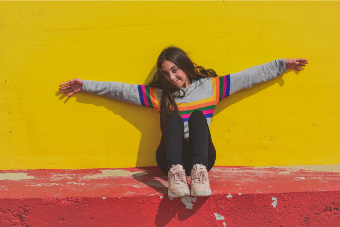 A tween or teen girl with long brown hair and a multicolored striped sweater sits against a yellow wall with her arms stretched out as she grows courage.