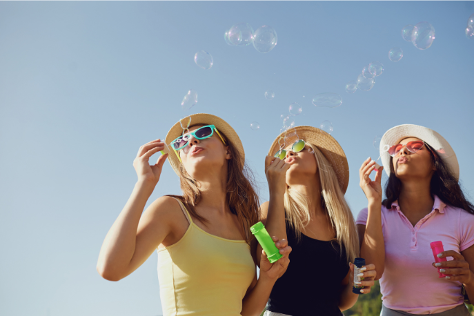 Three tween or teen girl friends with sunglasses and brimmed hats blow bubbles on a sunny day.