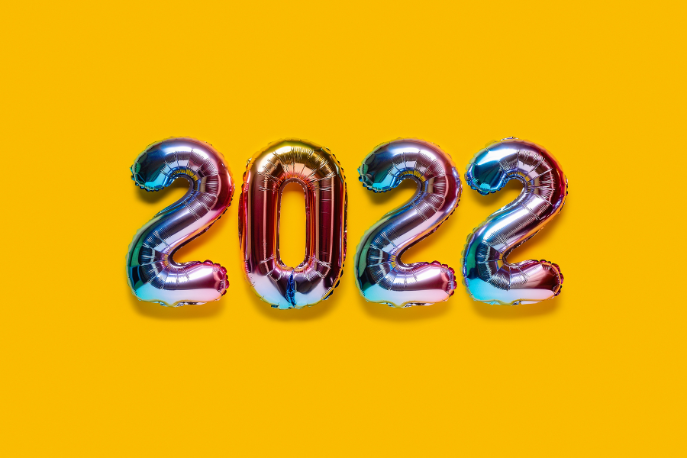 Metalllic multicolored mylar balloons spell out 2022 in front of a marigold yellow background.