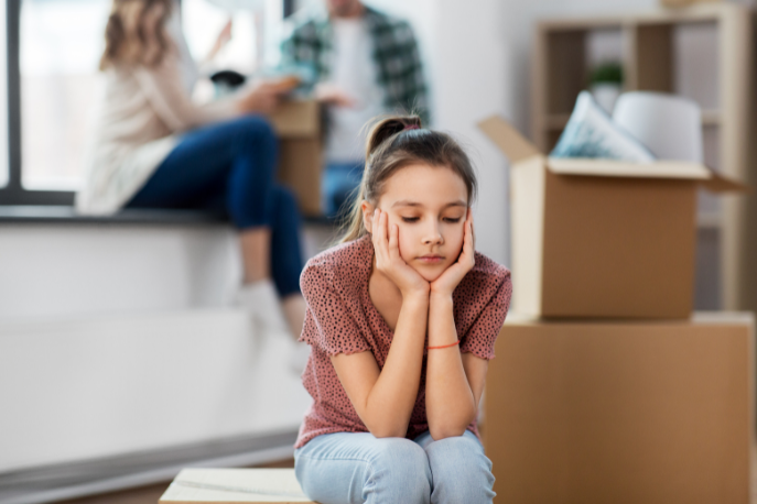 A tween or teen girl with a mauve top and light jeans sits amongst moving boxes.