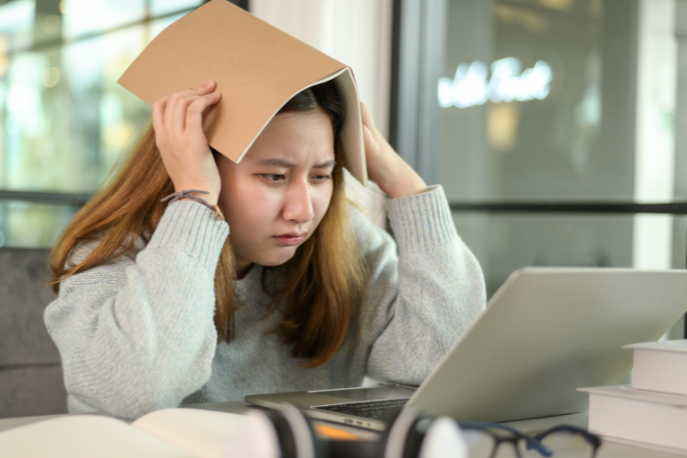 An asian tween or teen girl with red hair wearing a gray sweater sits at her computer with a notebook open on her head and looks stressed out.