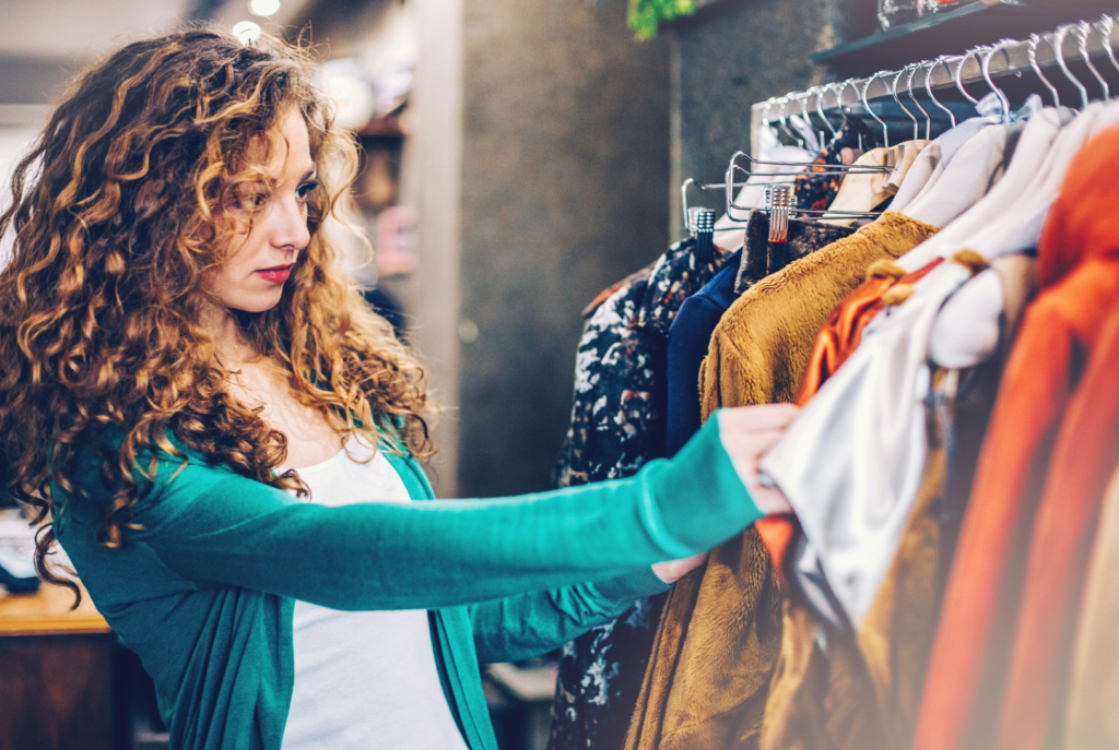 A tween or teen girl with curly auburn hair looks at clothes on a rack while shopping at a store.