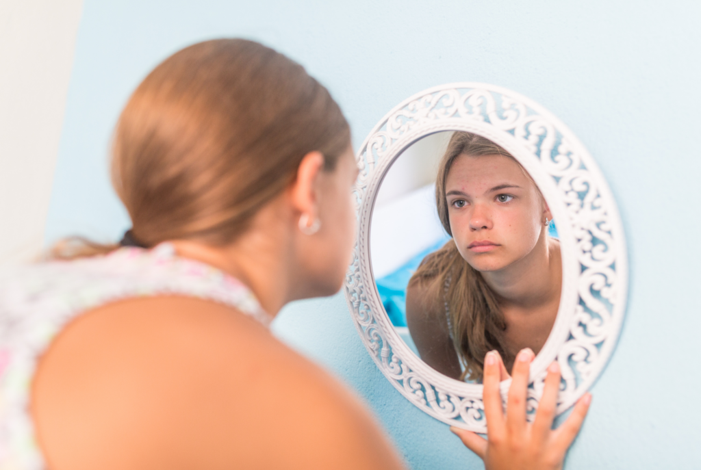 A tween or teen girl compares herself as she stares at her reflection in a mirror.
