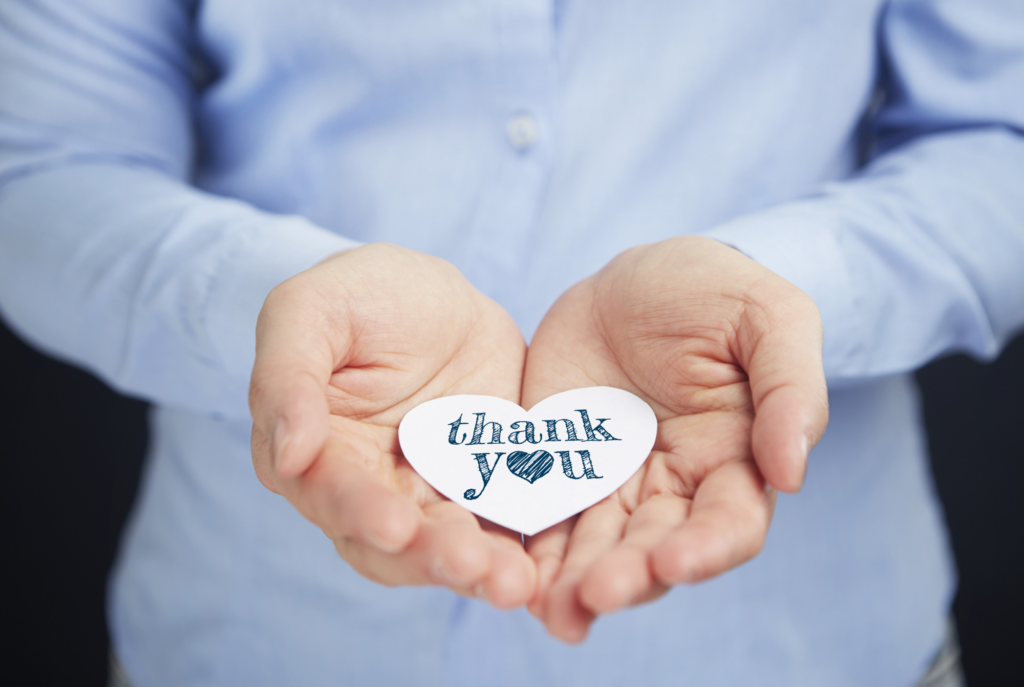 A tween or teen girl wearing a light blue button up shirt holds a heart-shaped thank you card in the open palms of her hands.
