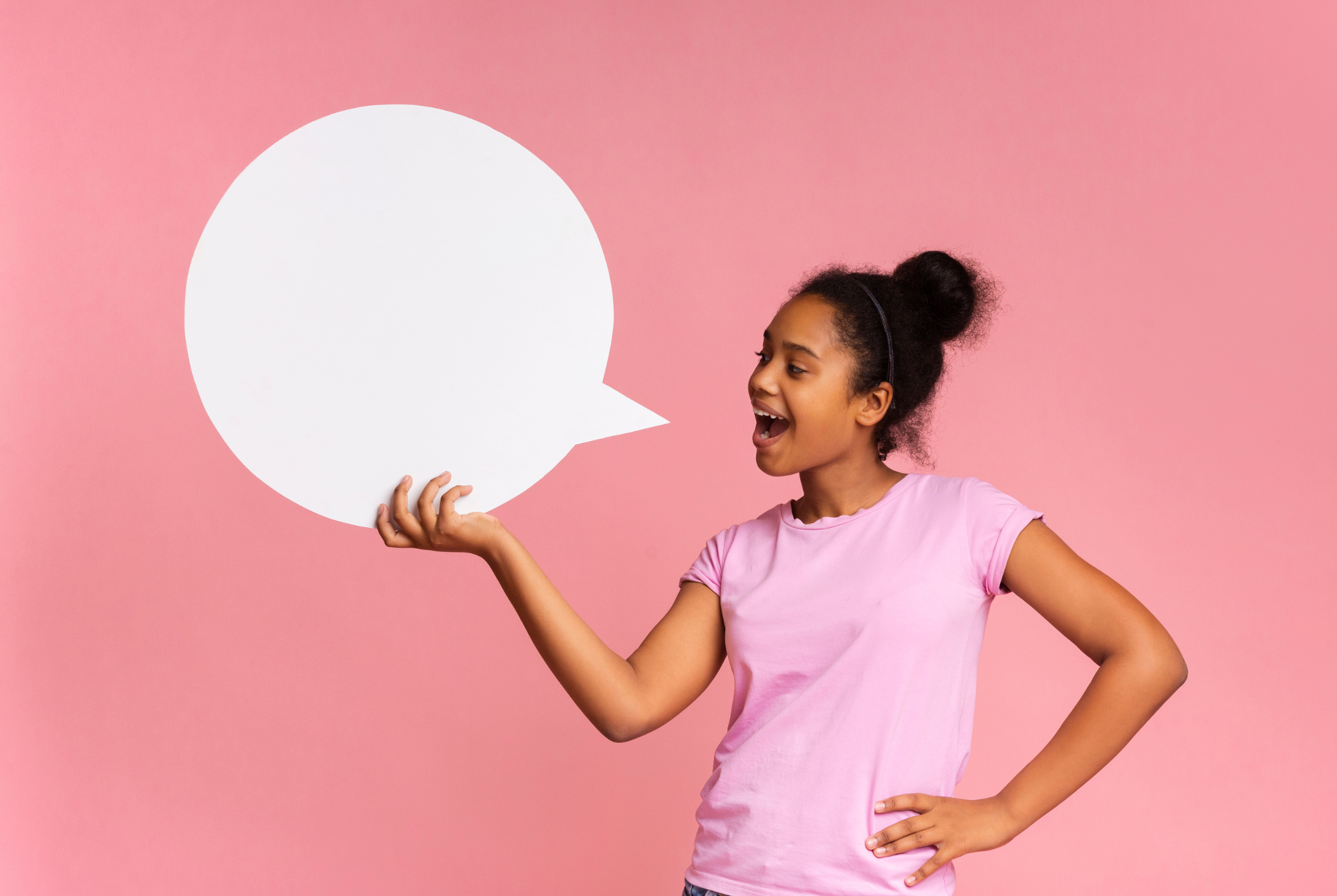 A tween or teen girl with brown skin and black hair wearing a light pink shirt holds a speech bubble as if she is about to say something.