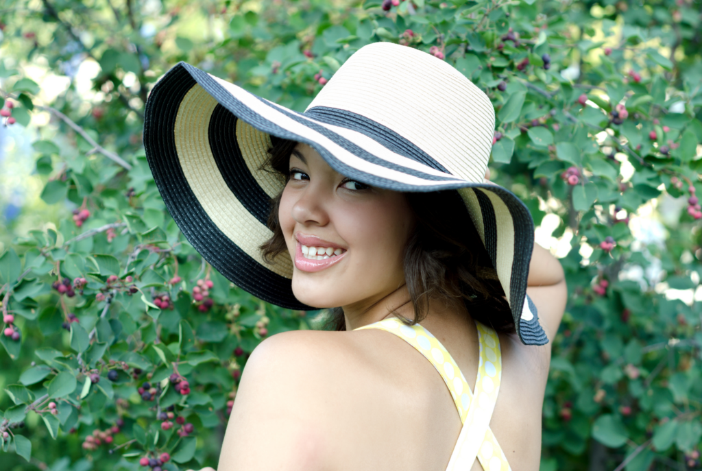 A tween or teen girl with tan skin and shorter black hair wearing a striped sunhat smiles while looking back and reflecting on the past year.