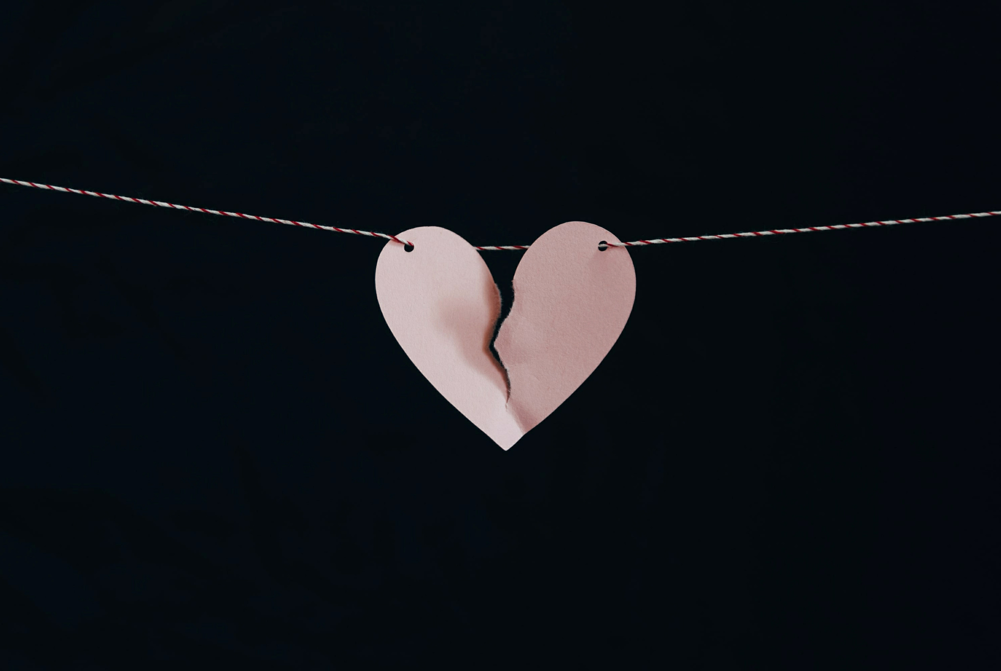A tween or teen girl's craft pink heart on a string hangs ripped in half against a black background.