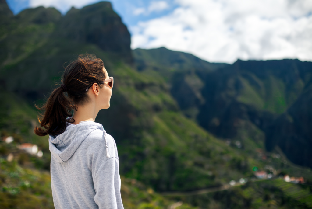 A tween or teen girl with brown hair wearing a gray hoodie and sunglasses looks ahead at a green mountainous valley, facing her future.