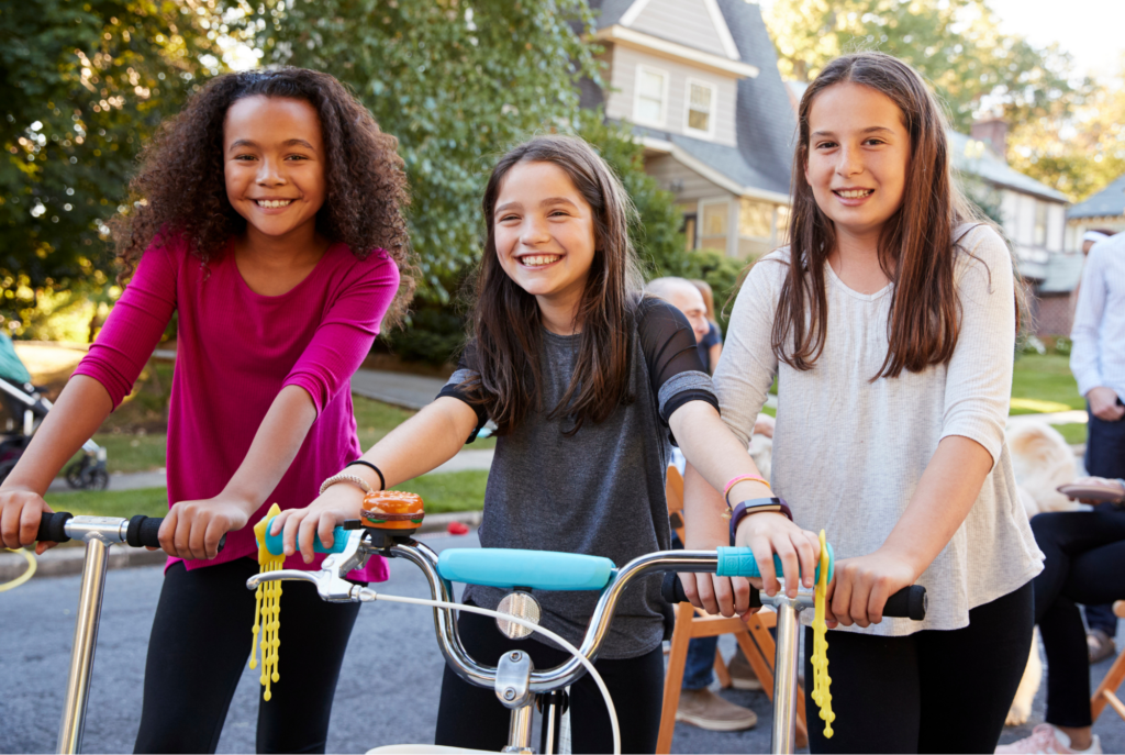 Tween or teen girls riding bikes and scooters outside smile at the camera.