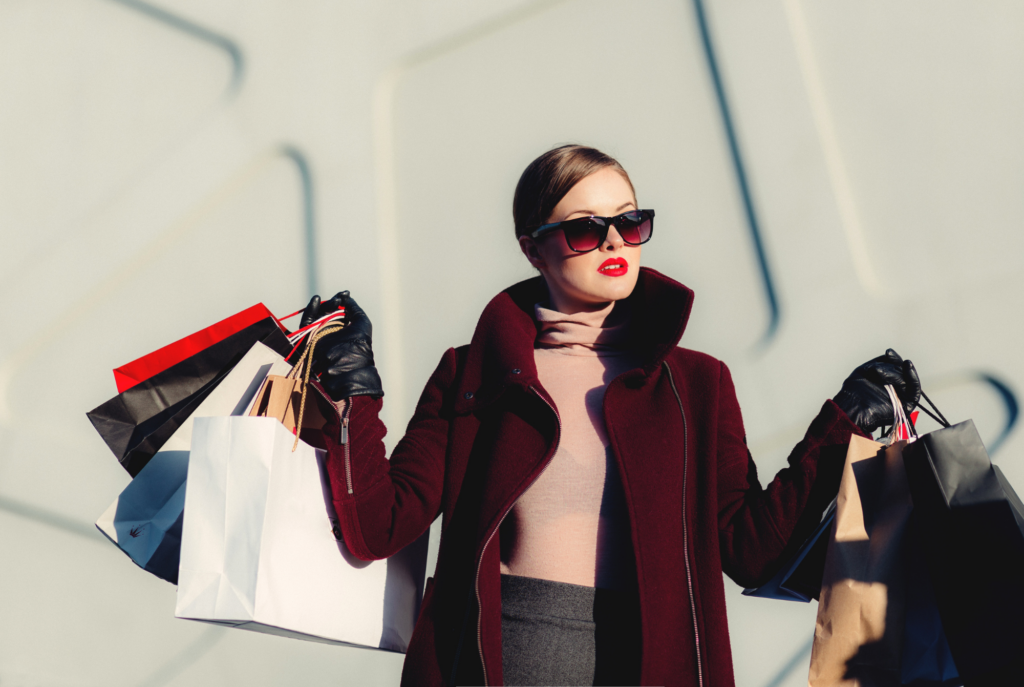 A tween or teen girl wearing slicked bun hair, sunglasses, red lipstick, and a stylish outfit holds up several shopping bags in both arms full of trendy items.