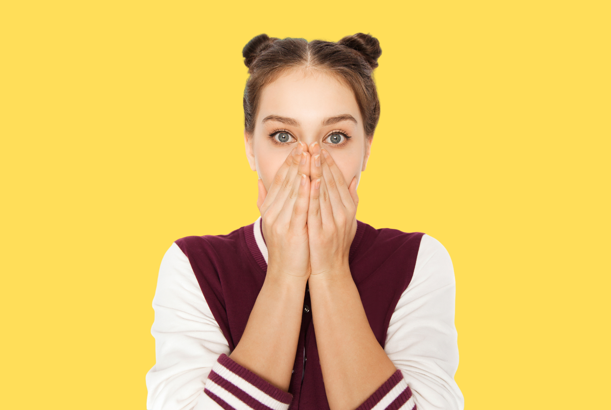 A tween or teen girl looks embarrassed as she stares straight into the camera with her hands over her mouth.