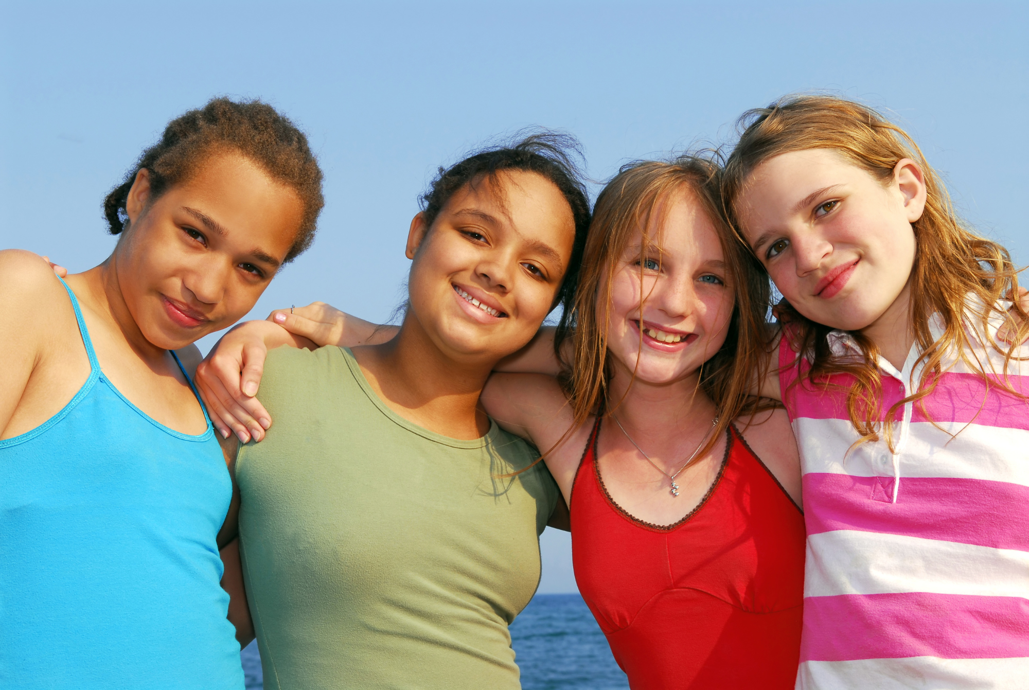 Four tween or teen girls wearing colorful tops stand with their arms around each other smiling in the sunlight because they know they are still enough.