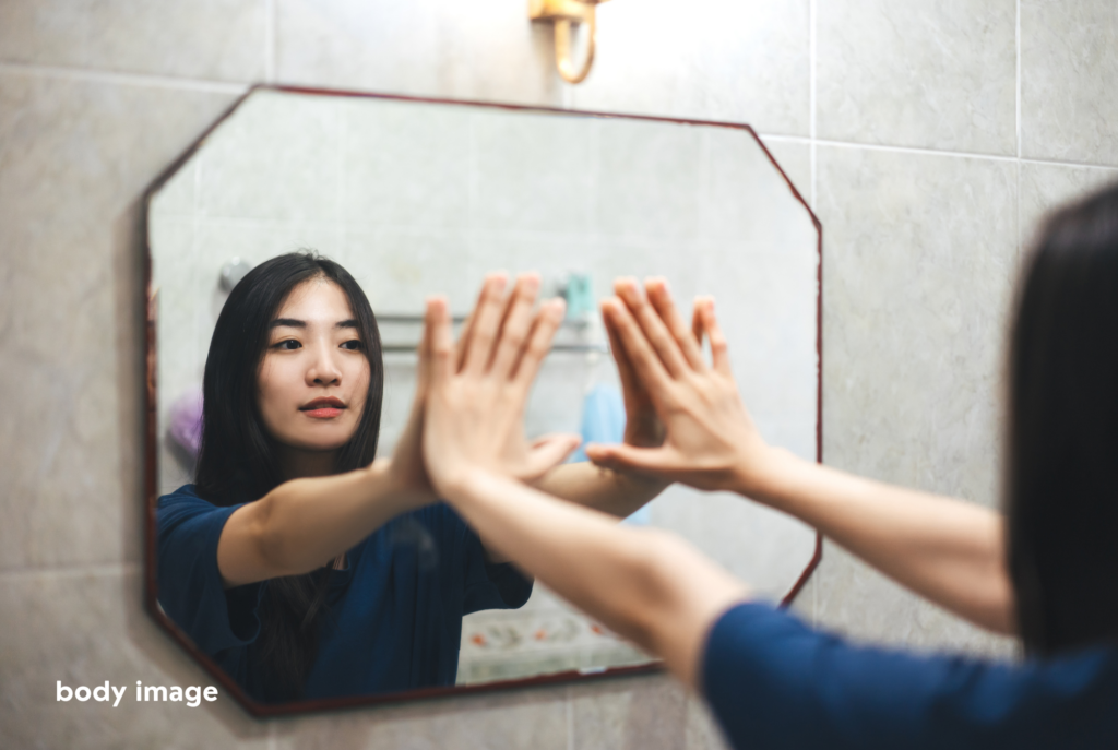 An asian teen or tween girl with long black hair stands in front of a mirror pressing her hands up against it