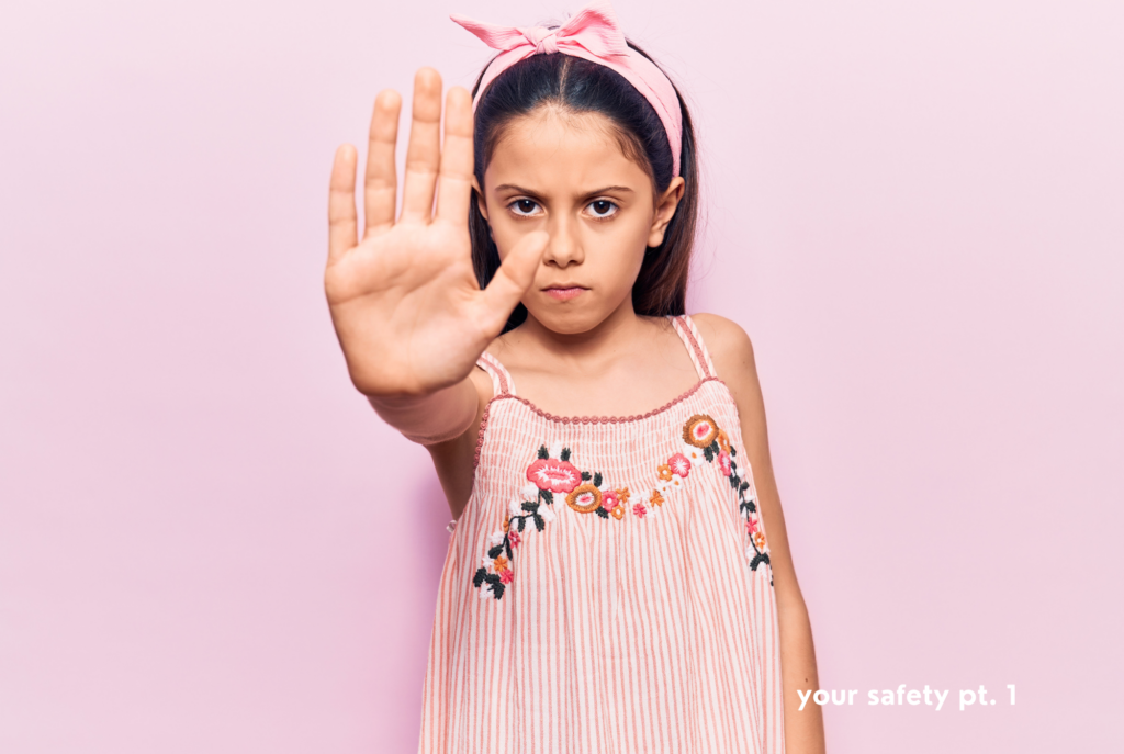 A tween or teen girl with light tan skin and long brown hair wearing a pink headband and dress stands in front of a light pink background with a serious expression and her arm extended in front of her and her hand signaling stop.