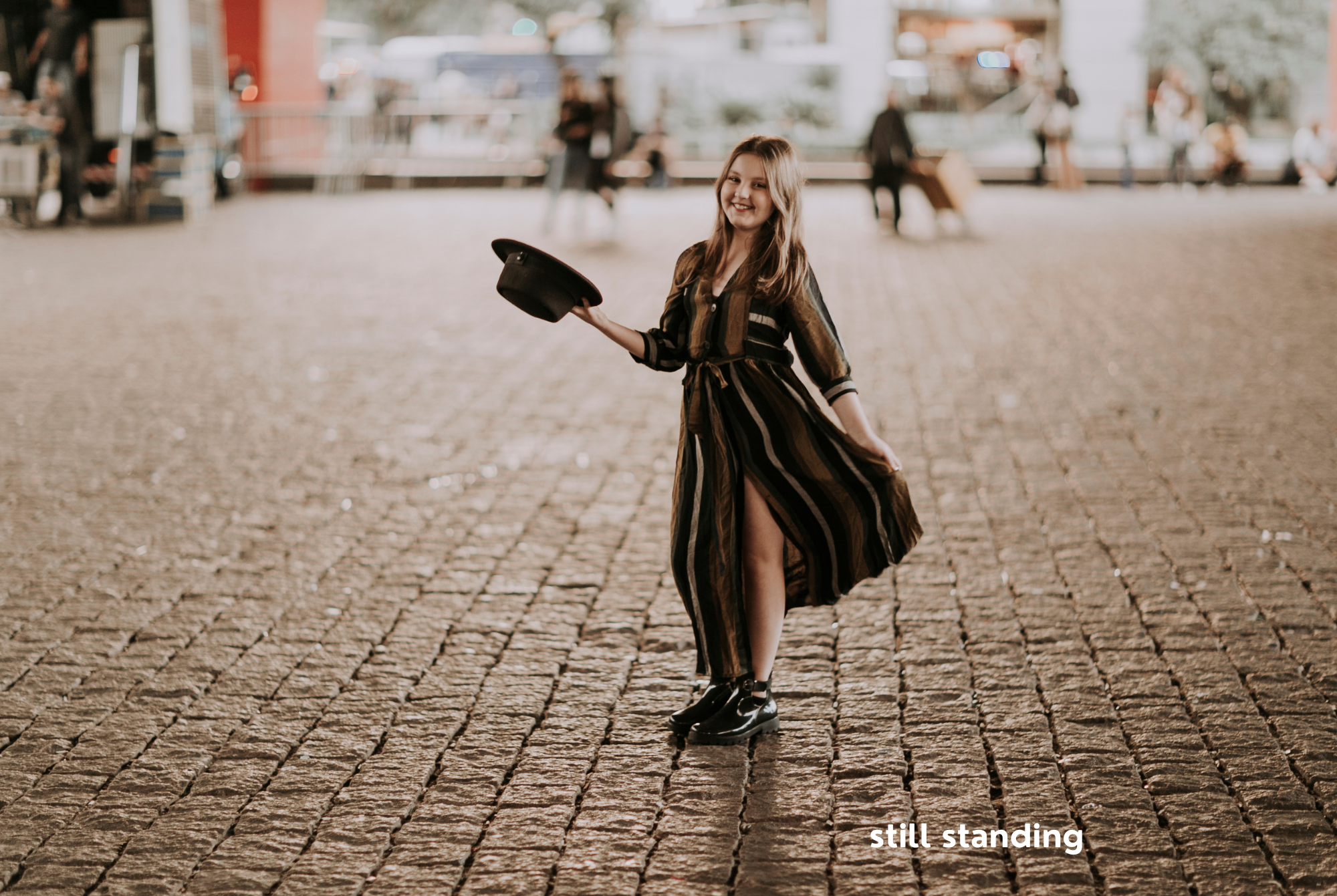 A tween or teen girl with long blonde hair and a long dress holding out a black hat stands on a cobblestone street at night.