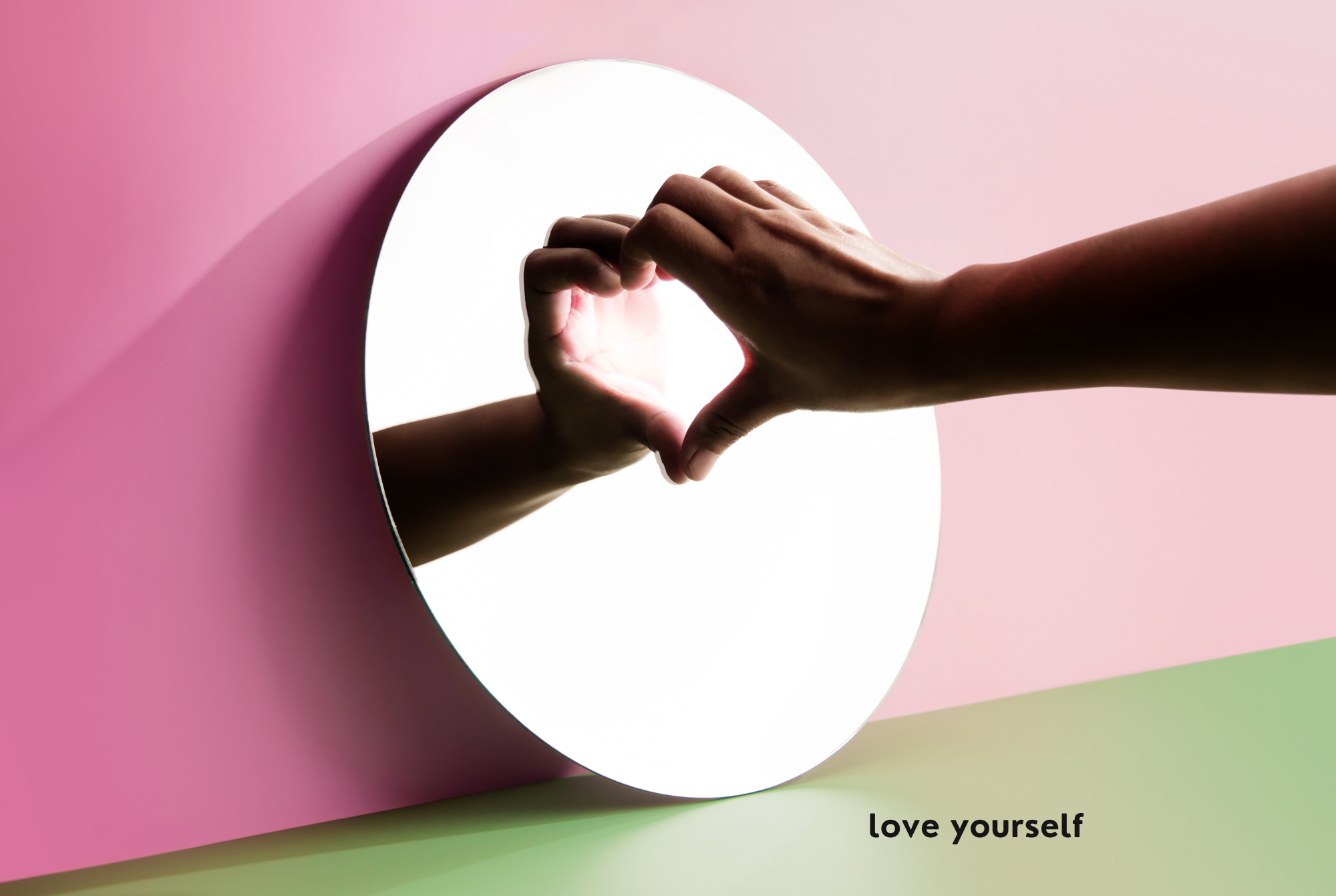 A tween or teen girl shows herself love as she holds her hand up to a round mirror, creating a heart shape with her hand's reflection.