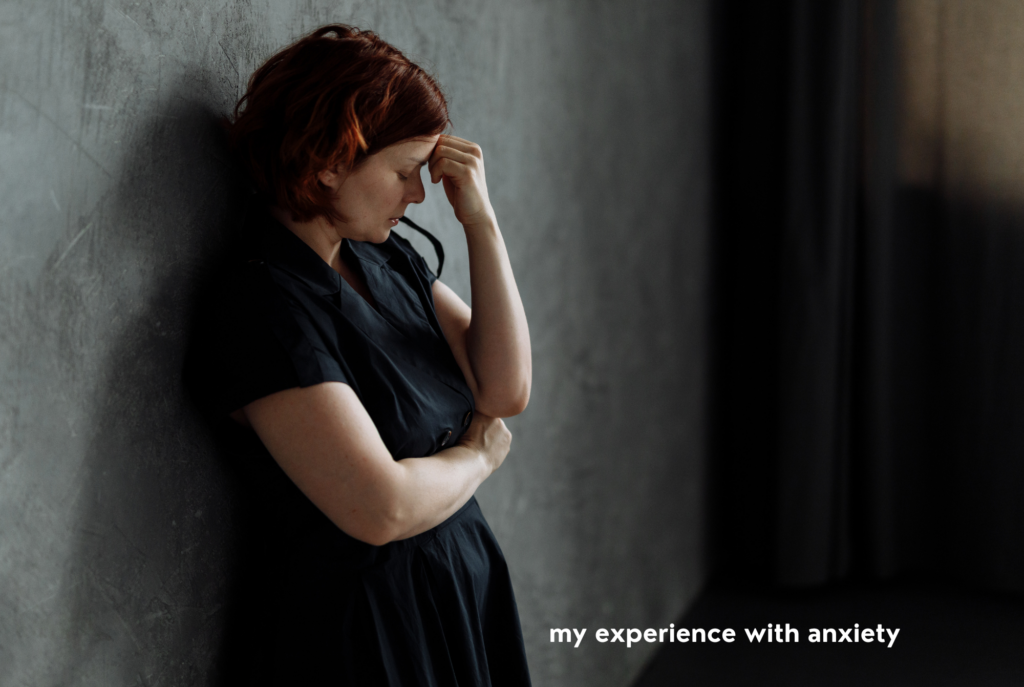 A teen or tween girl with red hair and a black dress leans against a gray concrete wall resting her head on her hand looking anxious.