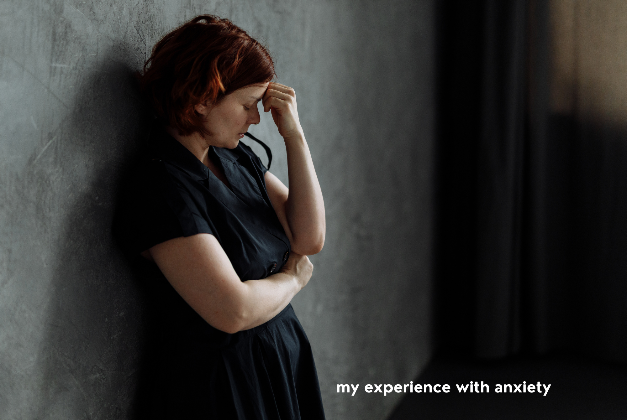 A teen or tween girl with red hair and a black dress leans against a gray concrete wall resting her head on her hand looking anxious.