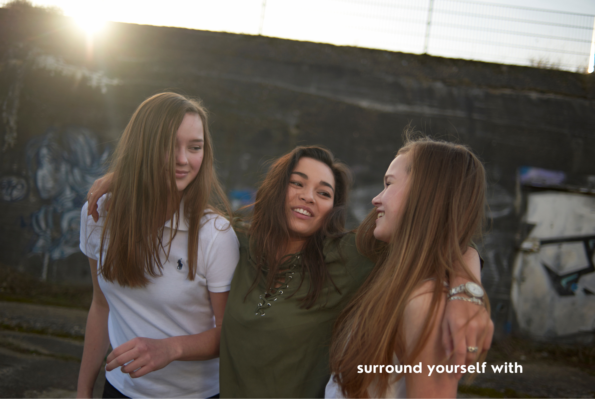 A tween or teen girl with tan skin and long dark brown hair walks outdoors surrounding herself with two friends with light brown and blonde hair.