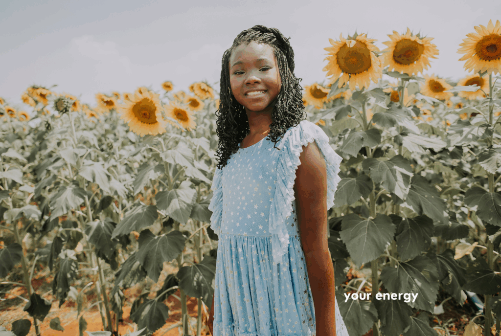 A tween or teen girl with dark skin and black braided hair wearing a light blue sundress stands next to tall sunflowers.