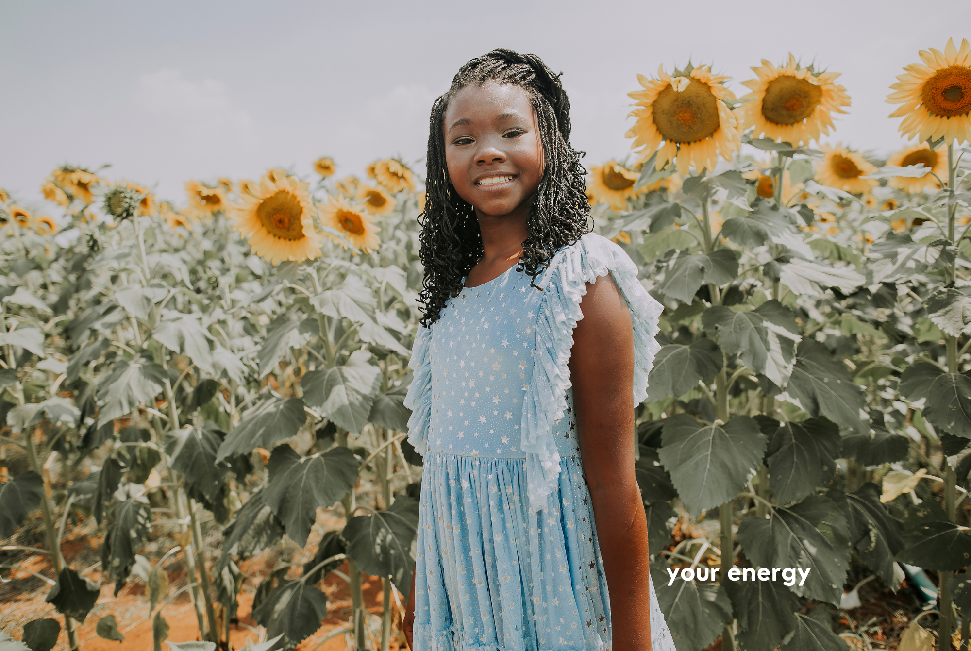 A tween or teen girl with dark skin and black braided hair wearing a light blue sundress stands next to tall sunflowers.