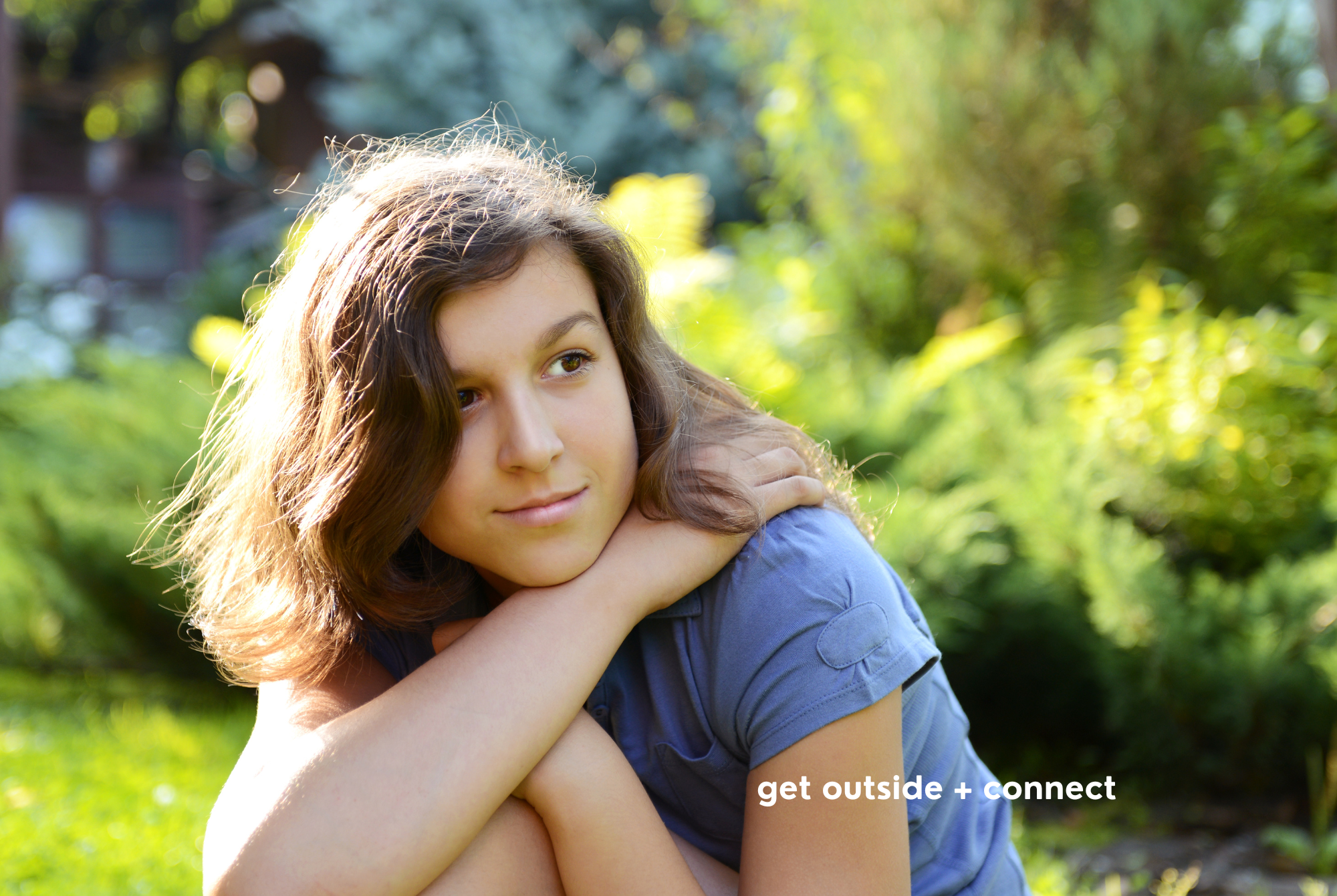 A teen or tween girl with tan skin and brown hair gets outside and connects with nature with grass, bushes, and sunlight.