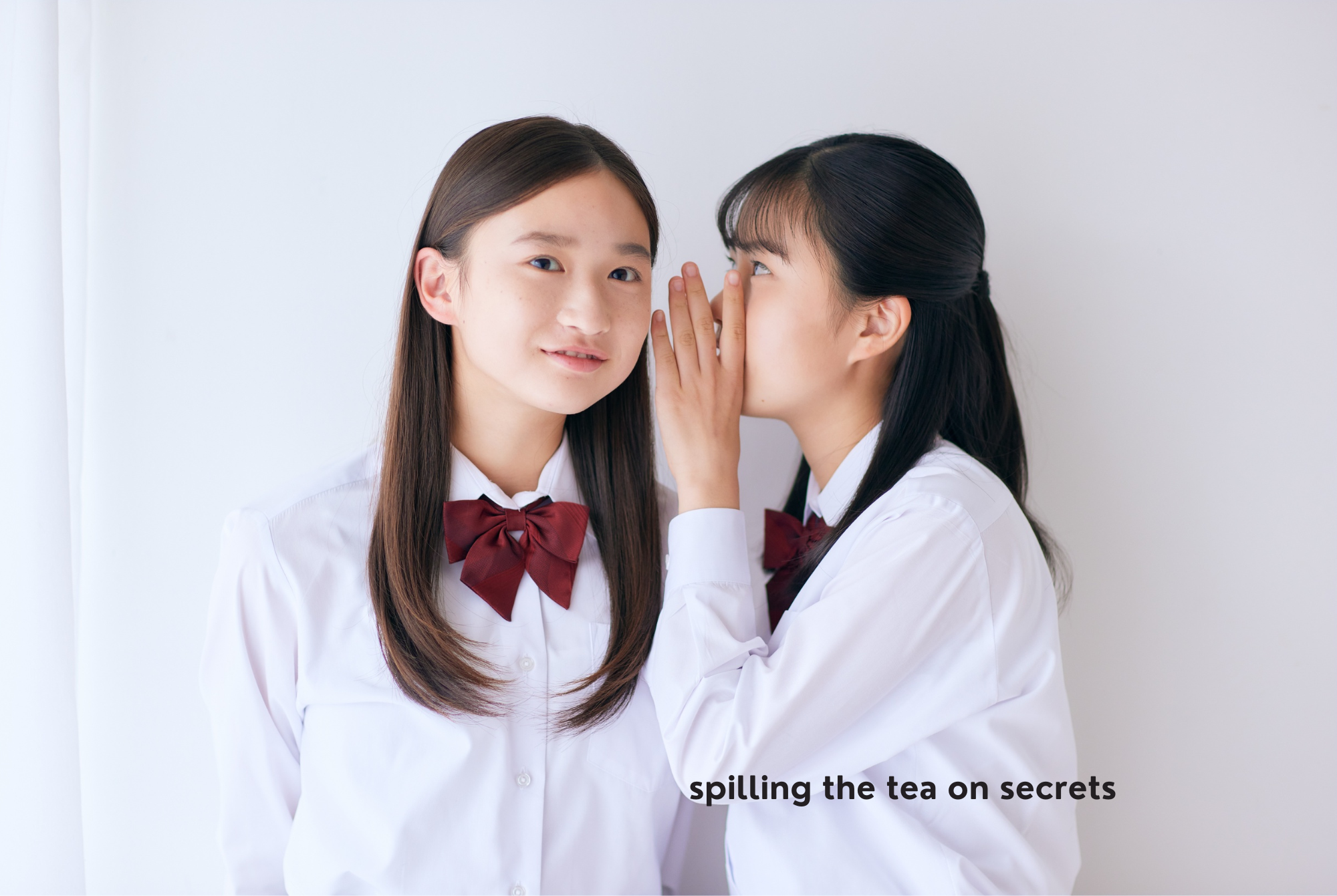 Two teen or tween girls with long, straight, brown hair wearing school uniforms share a secret privately.