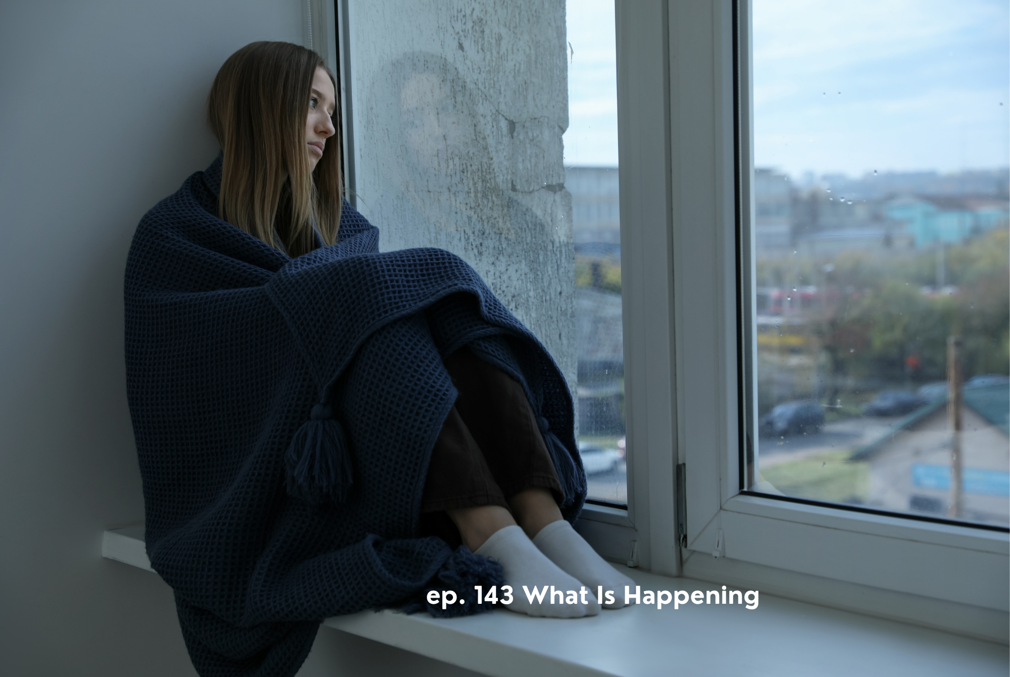 A teen or tween girl sits wrapped in a blanket next to a large window, feeling overwhelmed as she looks out at what is happening in the world.